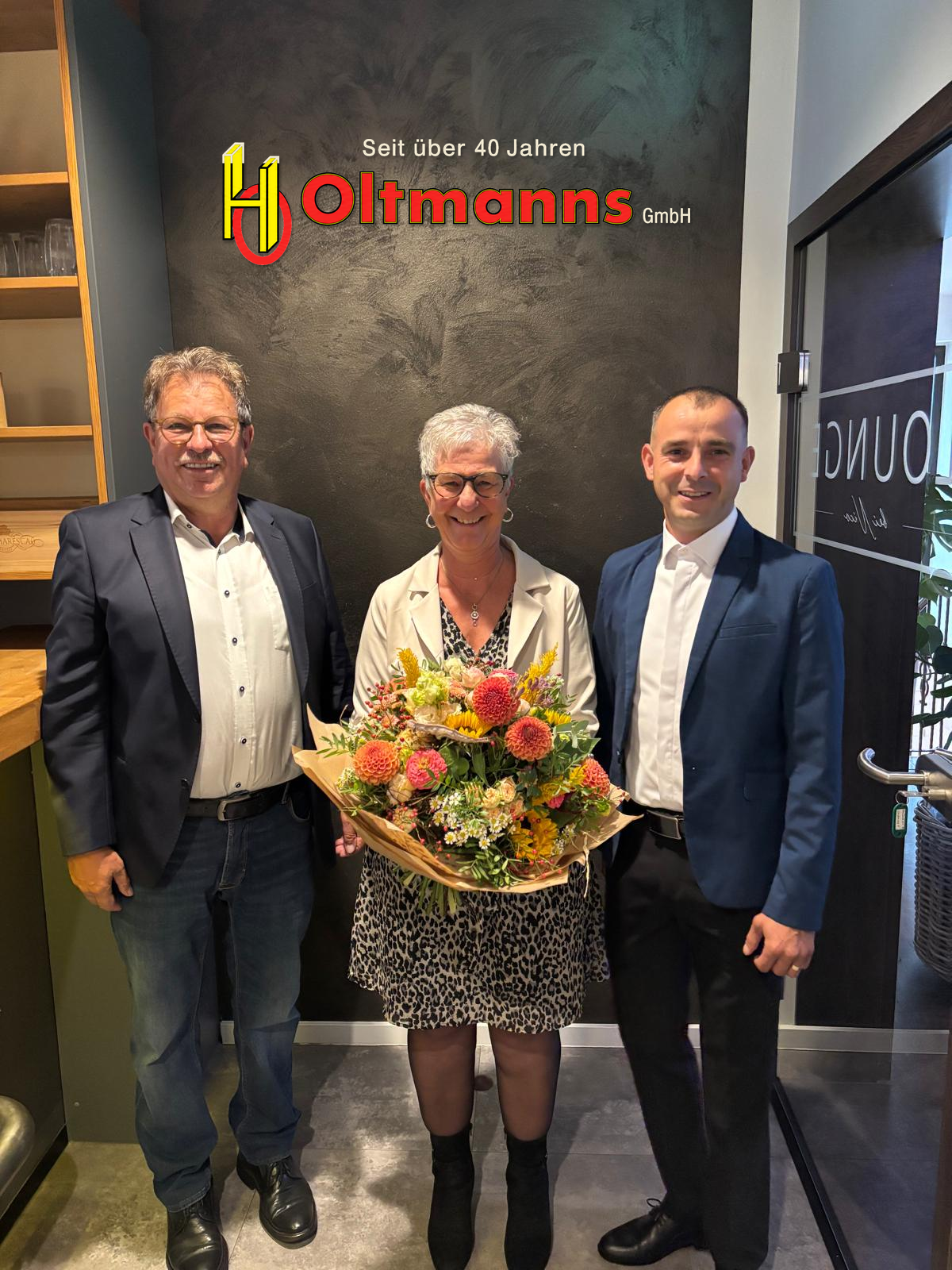 Gruppenfoto mit Blumenstrauß und Oltmanns-Logo im Hintergrund